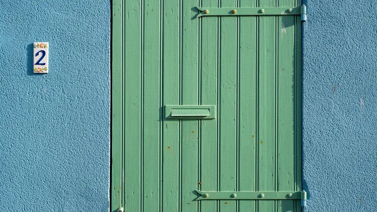 Detalle de una puerta de madera verde con dos bisagras metálicas de pletina a la vista y una ranura para el correo.