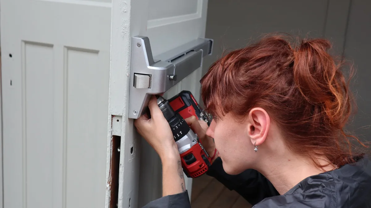 Una mujer taladra el canto de una puerta blanca para colocar la cerradura, con una plantilla metálica fijada a la puerta.