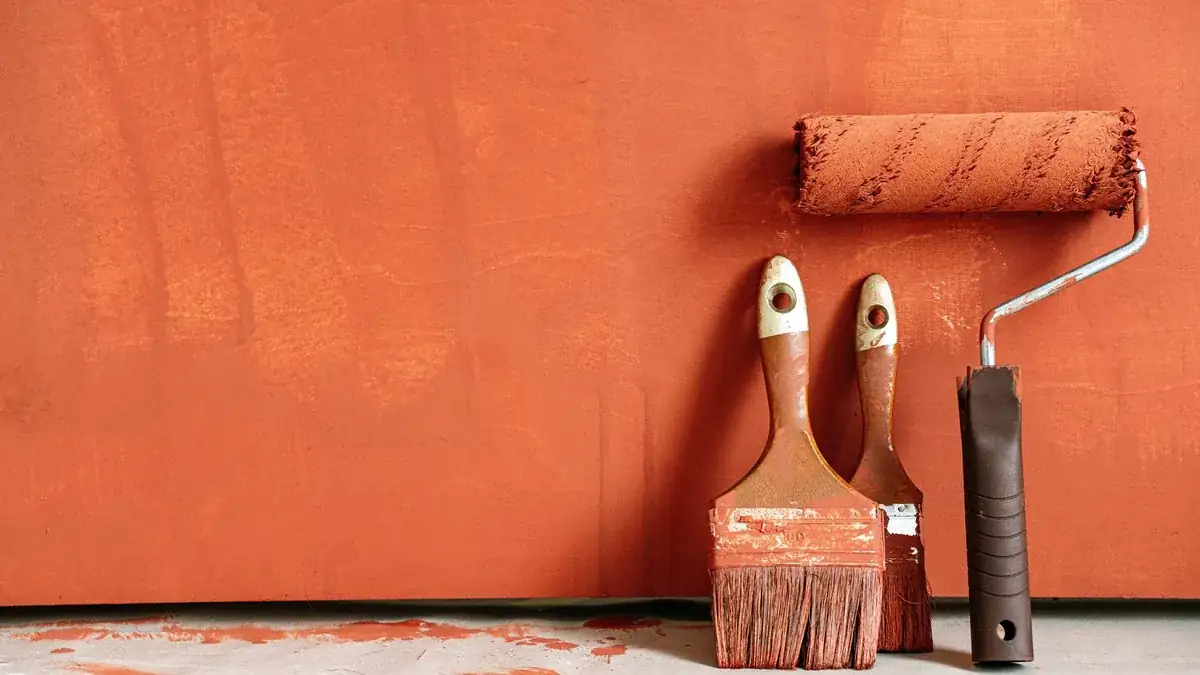 Dos brochas y un rodillo manchados de pintura naranja, apoyados contra una pared pintada del mismo color.