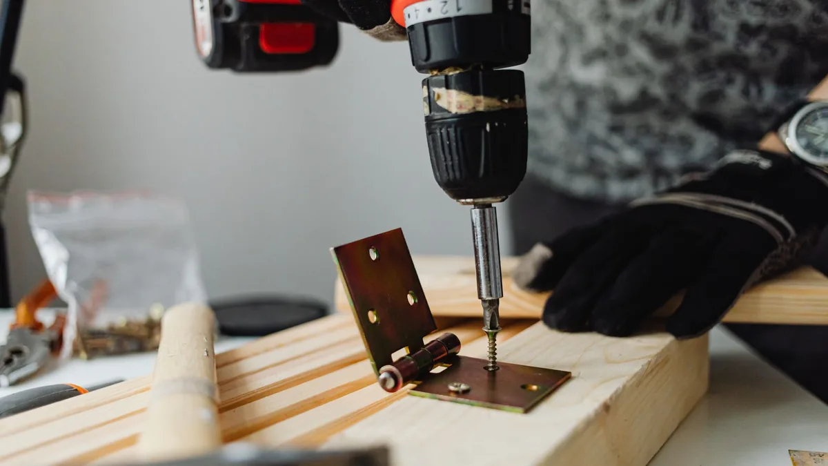 Unas manos con guantes atornillan con un taladro una bisagra metálica sobre un listón de madera.